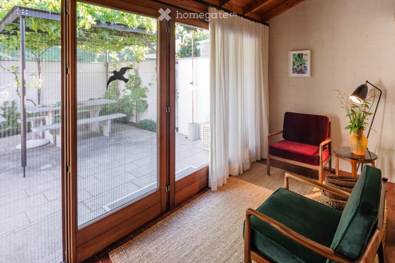 A cozy living room with a red sofa, green armchair, small round table, floor lamp, and a view of a garden through large glass doors.