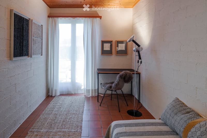 A modern, minimalist room with exposed brick walls, a wooden ceiling, and a large window with white curtains. The room features a sofa, a desk with a chair, a floor lamp, and several picture frames on the walls.