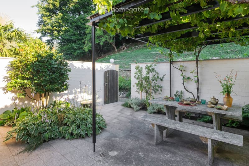 A well-maintained garden area with a stone table and benches, surrounded by lush greenery and a pergola with climbing plants. There is a small gate and a variety of plants and flowers.