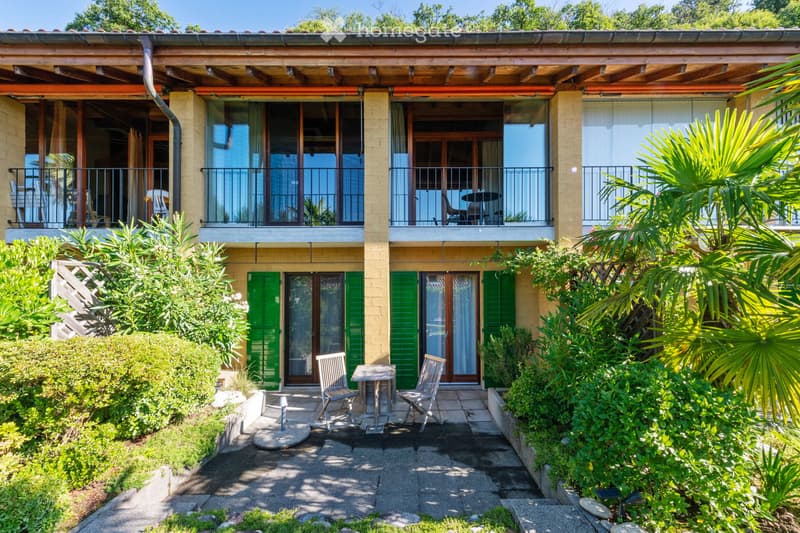 Two-story house with light yellow walls, green shutters, and large windows. Balconies on the upper level have outdoor furniture, and the ground level features a patio with a small table and chairs surrounded by lush greenery and palm trees.