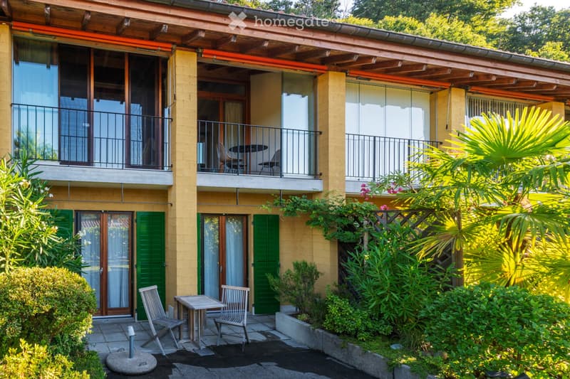 Two-story building with yellow walls, large windows, and balconies. Outdoor seating areas with tables and chairs are visible on the ground and upper levels, surrounded by lush greenery and palm trees.