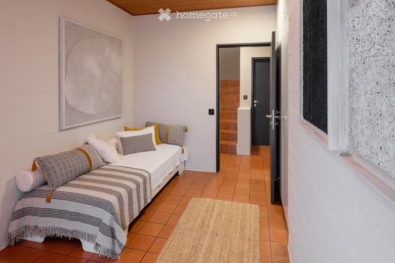 Cozy bedroom with a white sofa bed, decorative pillows, and a patterned blanket. The room features exposed brick walls, terracotta tile flooring, and a small rug near the bed. A doorway leads to a staircase.