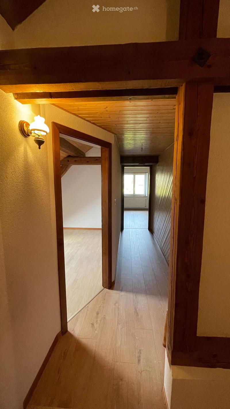 Wooden-floored hallway with warm lighting, open doorways leading to other rooms, and a visible window at the end.