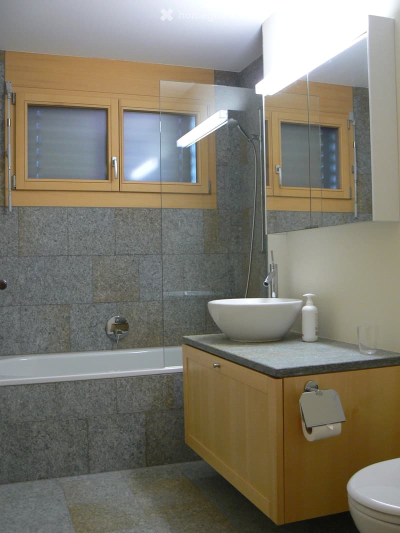 gray tile wall, white bathtub, white sink, wooden cabinet, toilet, shower head