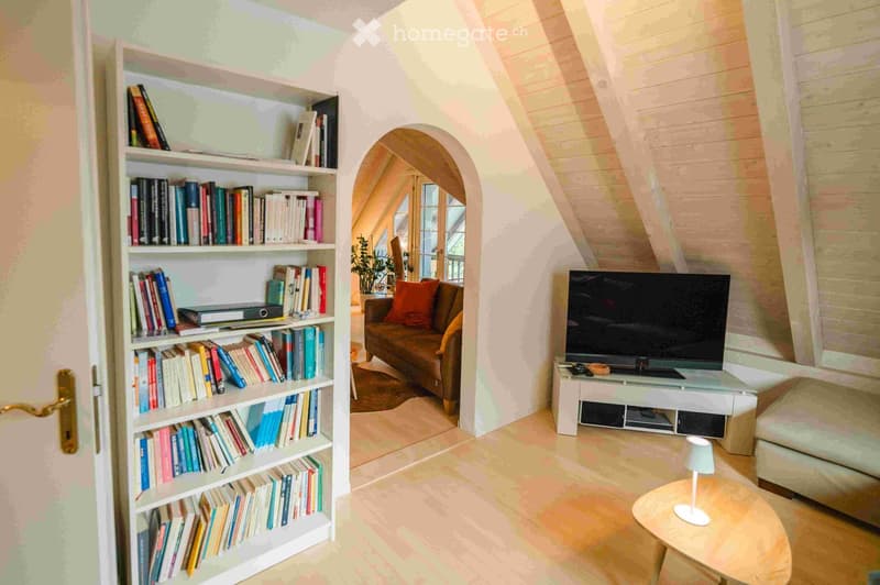 A cozy living room with a bookshelf filled with books, a sofa with orange cushions, a television on a stand, a small round table with a lamp, and a potted plant visible through an arched doorway.