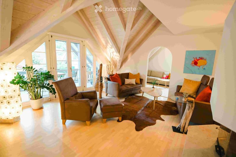 Bright living room with exposed wooden beams, large windows, and a mix of seating including a sofa, armchair, and ottoman. Features a coffee table, glass table, decorative plant, and artwork on the wall.