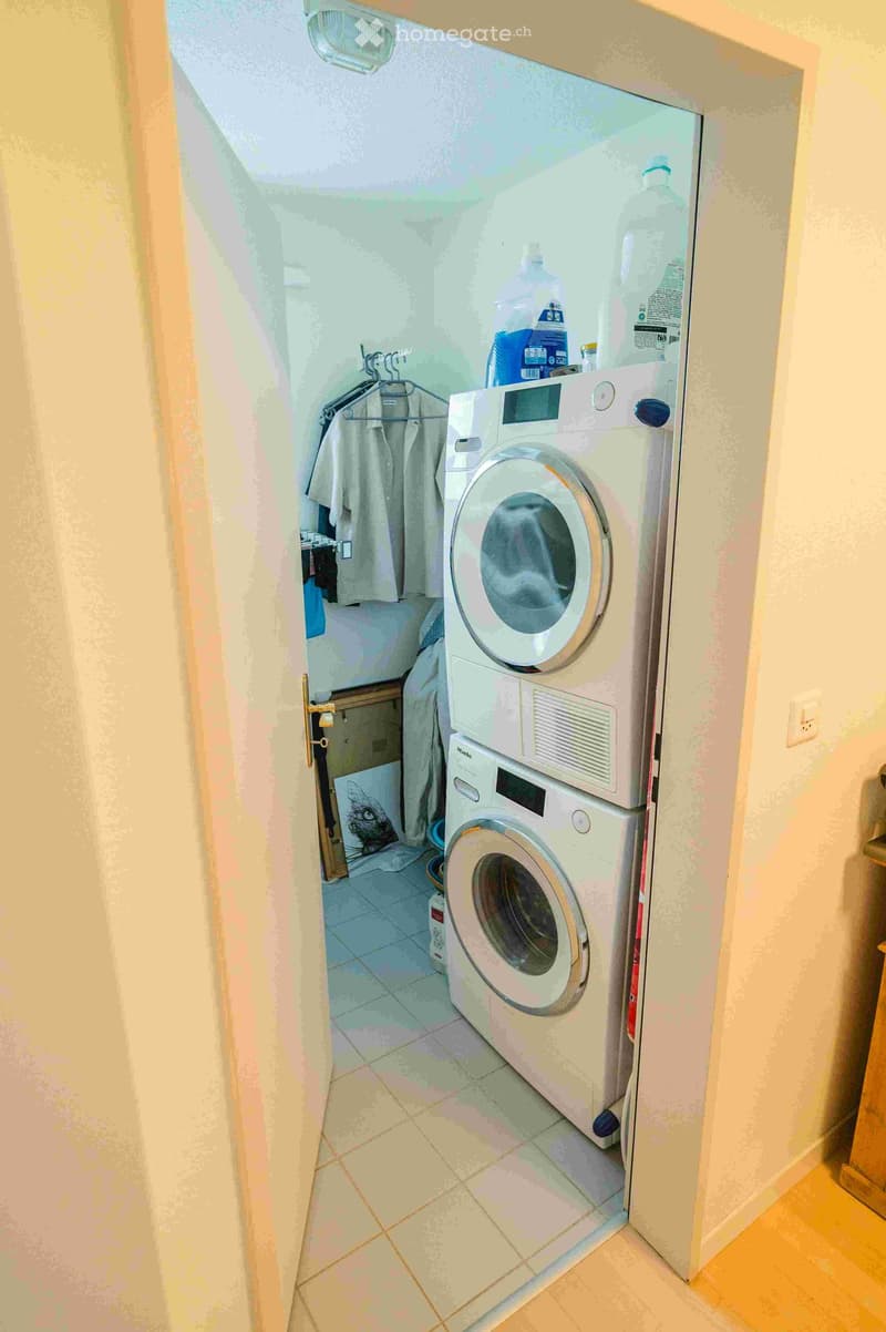 A small bathroom with a stacked washing machine and dryer, a towel hanging on a rack, and cleaning supplies on top of the dryer.