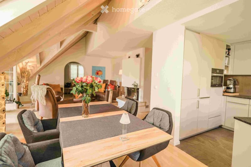 A modern, open-concept living and dining area with a grey sofa, wooden dining table, chairs, a lamp, and a vase of flowers. The kitchen area features white cabinets and stainless steel appliances.