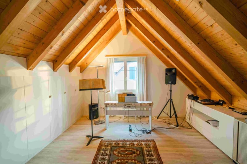 An attic room with wooden beams and slanted ceilings, featuring a piano, speakers, a tripod, a table with a keyboard, and a cabinet.