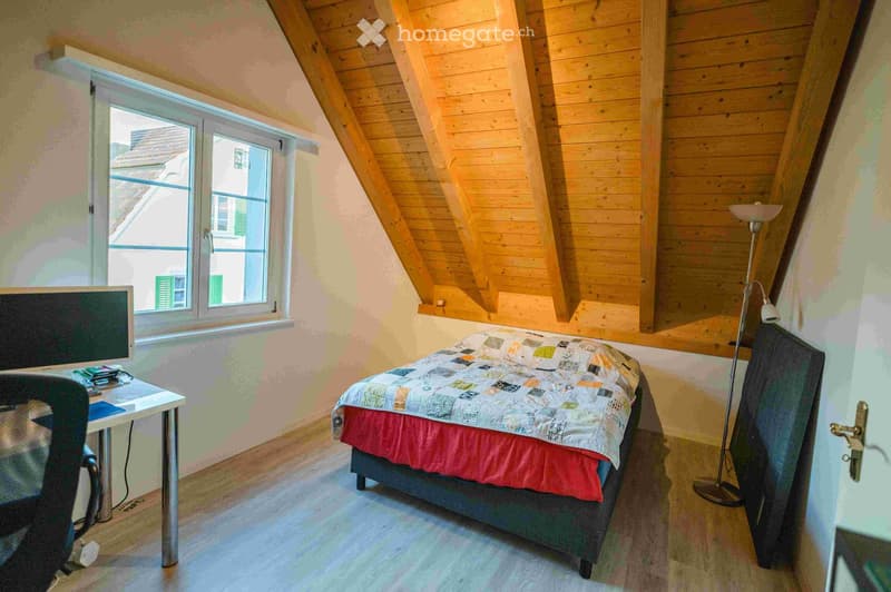 Cozy attic bedroom with wooden slanted ceilings, a double bed with a colorful quilt, a desk with a computer, and a floor lamp near the window.