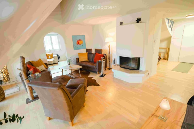 Modern living room with a fireplace, large windows, and comfortable seating including a sofa, armchair, and ottoman. The space features a coffee table, floor lamp, and decorative elements like a painting and plants.