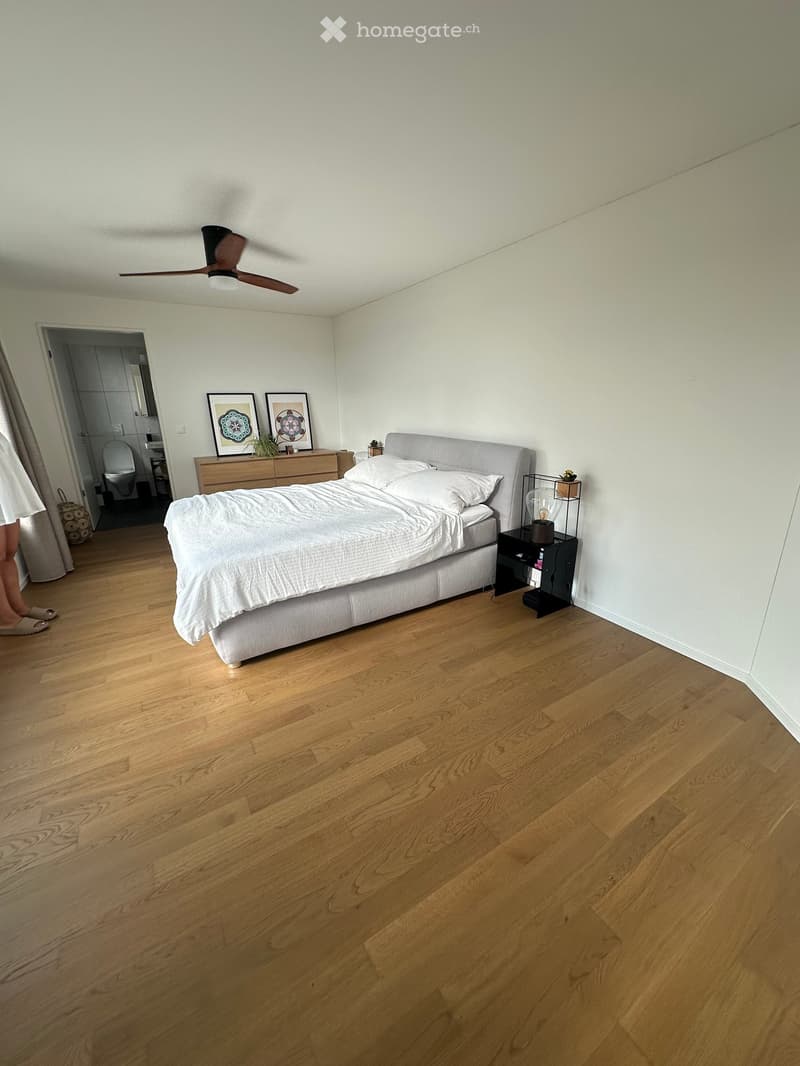 Spacious bedroom with wooden flooring, a large bed with white bedding, a nightstand with a lamp, a dresser with framed artwork, and a ceiling fan.