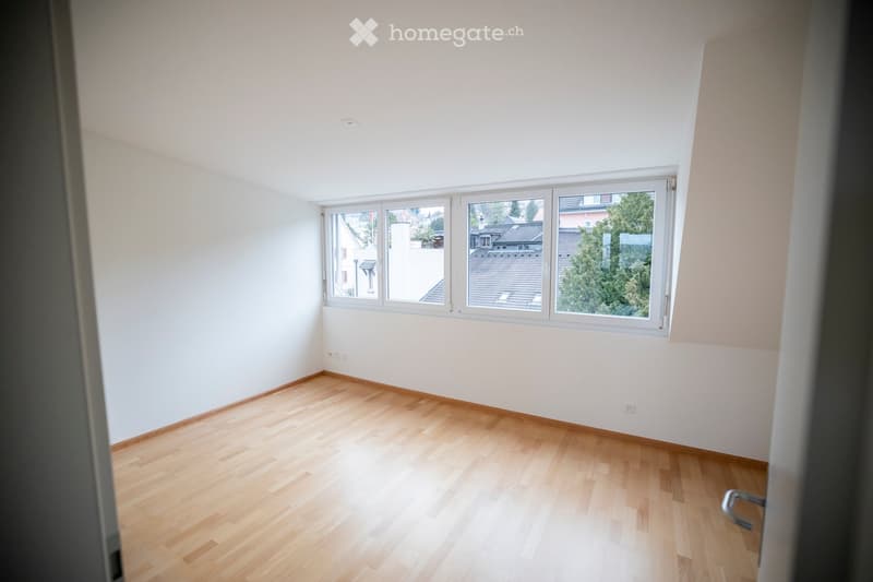 Empty room with light wooden flooring and white walls, featuring large windows offering a view of rooftops and trees.