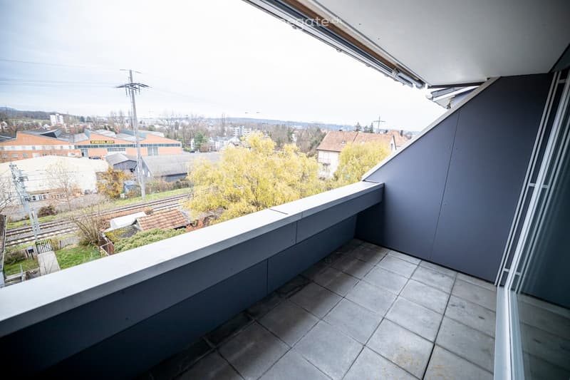 Modern balcony with tiled floor, white railing, and expansive city view including buildings, trees, and railway tracks.