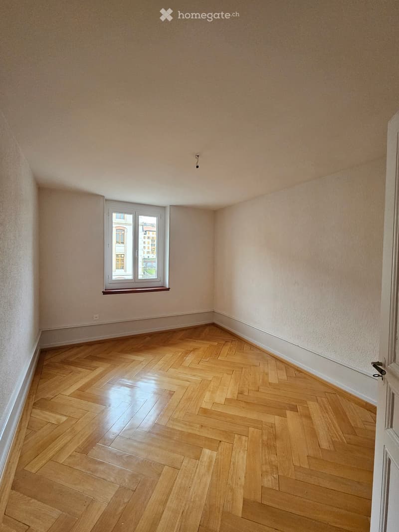 Empty room with light-colored walls and polished wooden herringbone flooring, featuring a single window with a small sill and a door frame visible on the right side.