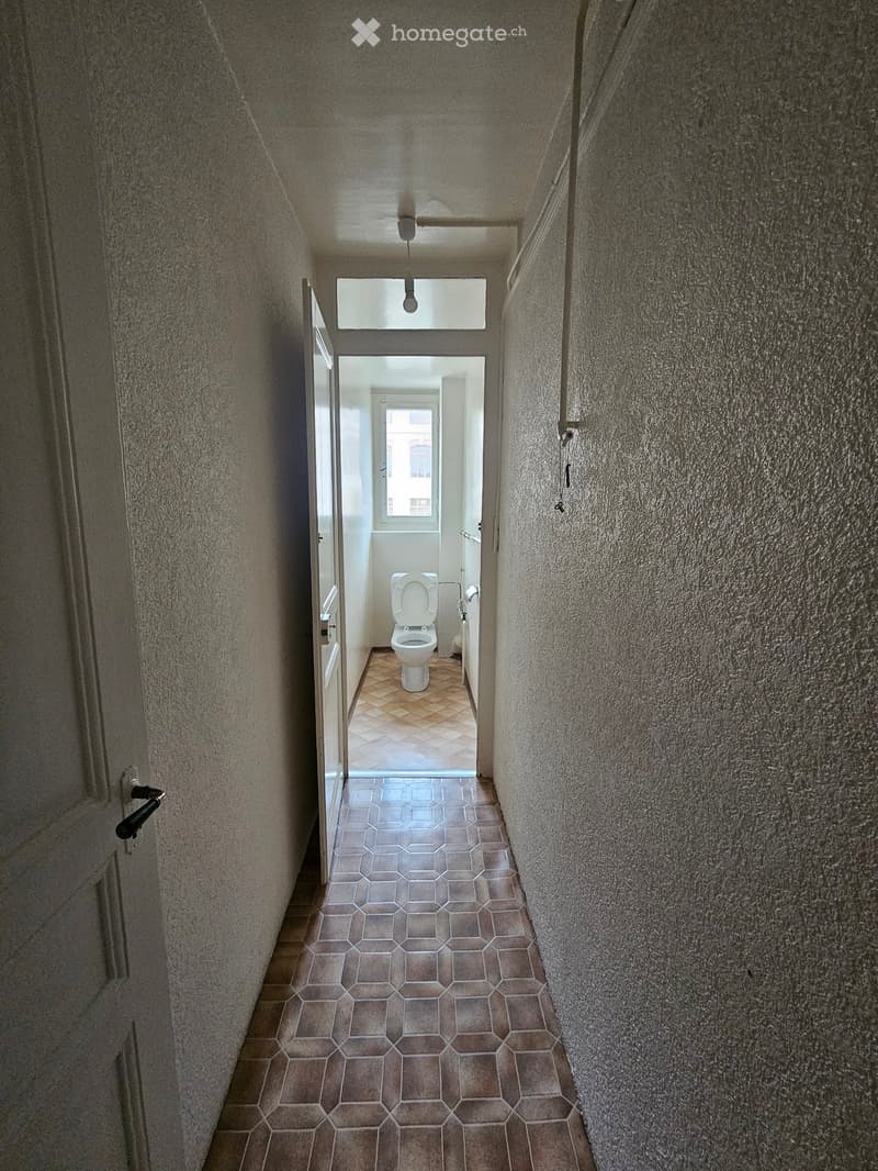 A narrow hallway with tiled flooring and textured walls, leading to a bathroom with a toilet and a window.