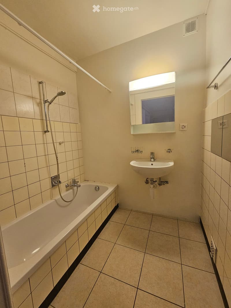 Modern bathroom with a bathtub, sink, mirror, and showerhead. Light-colored tiles and a sleek design.