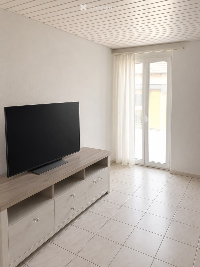 Modern living room with a large flat-screen TV on a wooden TV stand, white tiled floor, and large sliding glass doors with white curtains.