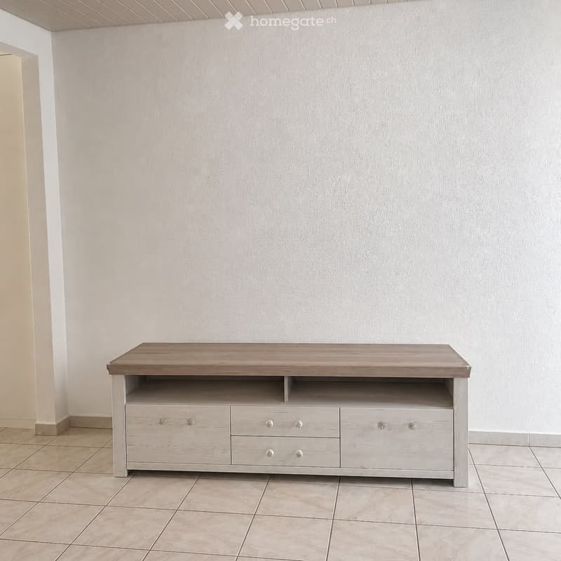 Empty room with light-colored walls and tiled flooring, featuring a modern TV stand with drawers and a shelf compartment.