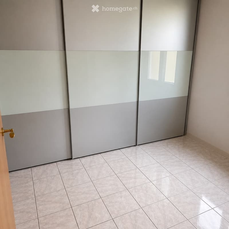 Empty room with beige tiled flooring and a large, modern sliding wardrobe with grey and white panels.