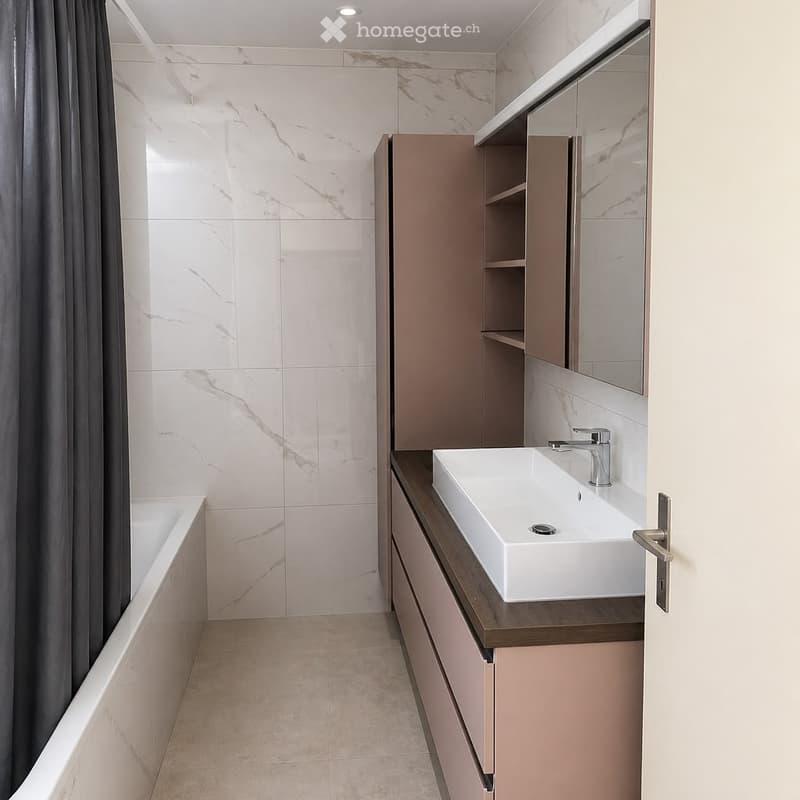 Modern bathroom with a white sink, chrome faucet, beige cabinet, and a large mirror above the sink. The walls are tiled in a marble pattern, and a bathtub is visible to the left.