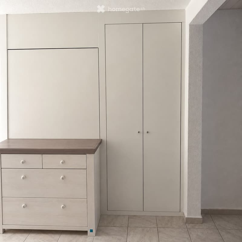 Empty room with light grey walls, tiled floor, built-in white wardrobes, and a small wooden dresser with drawers.