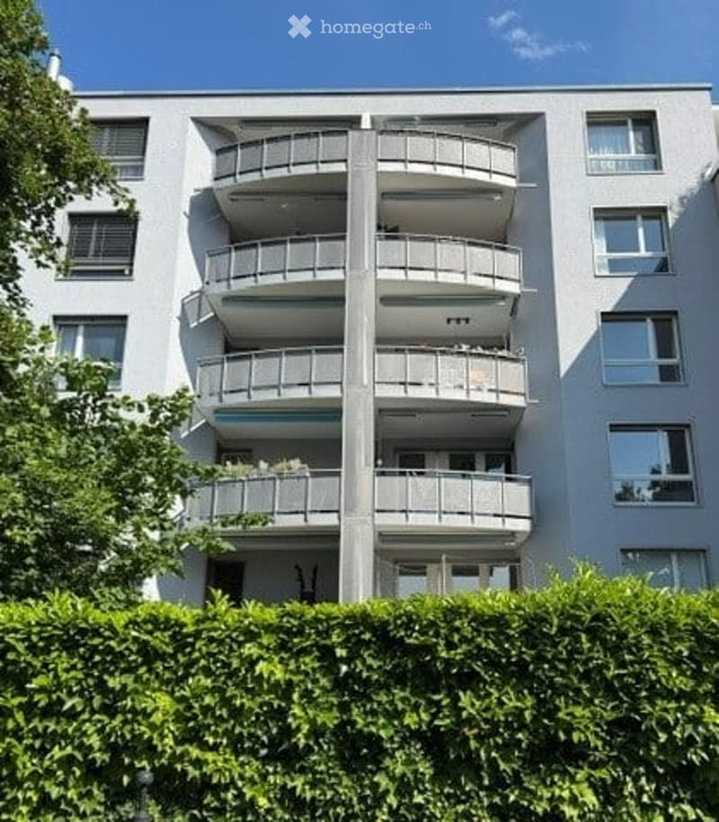 Modern multi-story apartment building with multiple balconies and large windows, surrounded by greenery.