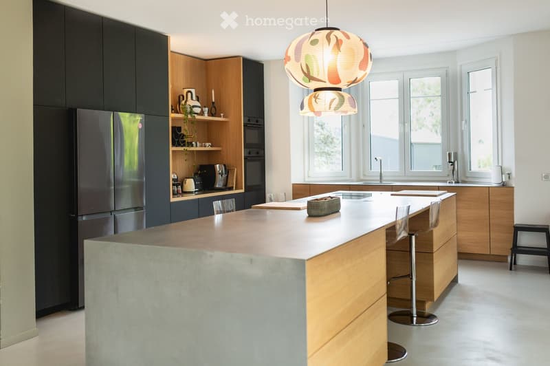 Modern kitchen with dark and light wood cabinets, stainless steel appliances, a large island with a concrete countertop, and a hanging lamp with a floral design.