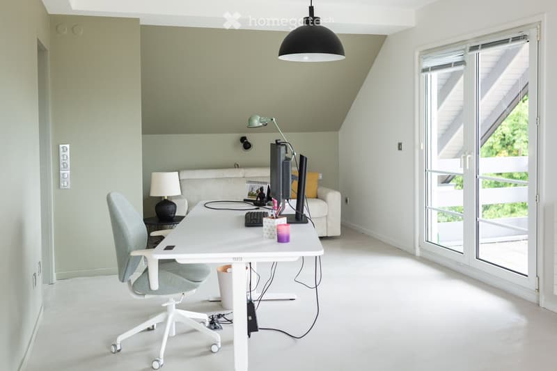 Bright, modern workspace with a white desk, ergonomic chair, computer setup, and a comfortable sofa area. Large windows and a hanging pendant light enhance the space.