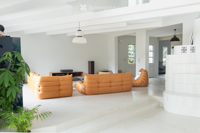 Modern living room with white walls, light-colored flooring, and large windows. Features a tan leather sofa, coffee table, pendant lights, and a speaker system.