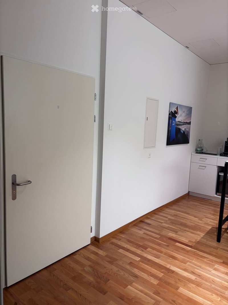 An empty room with white walls, wooden floor, and a closed door. There is a picture frame and power outlet on the wall, and a small table with a bottle on the right side.
