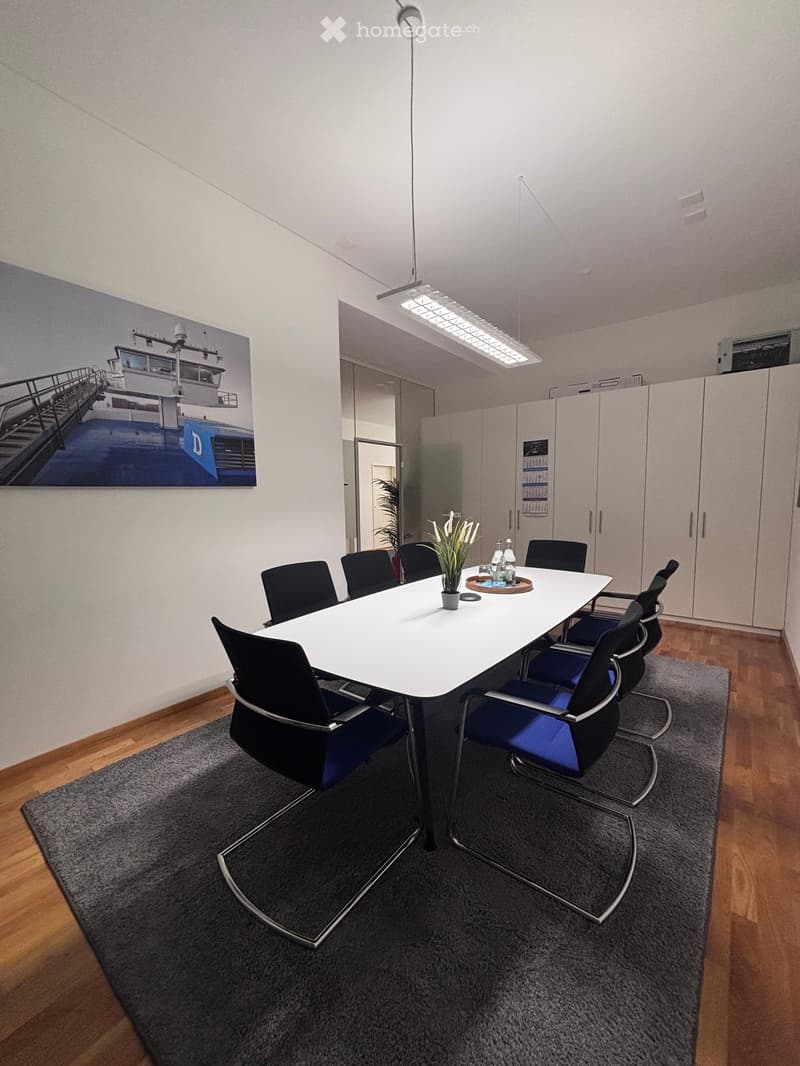 A conference room with a long white table, black and blue chairs, a vase with flowers, and a potted plant on the table.
