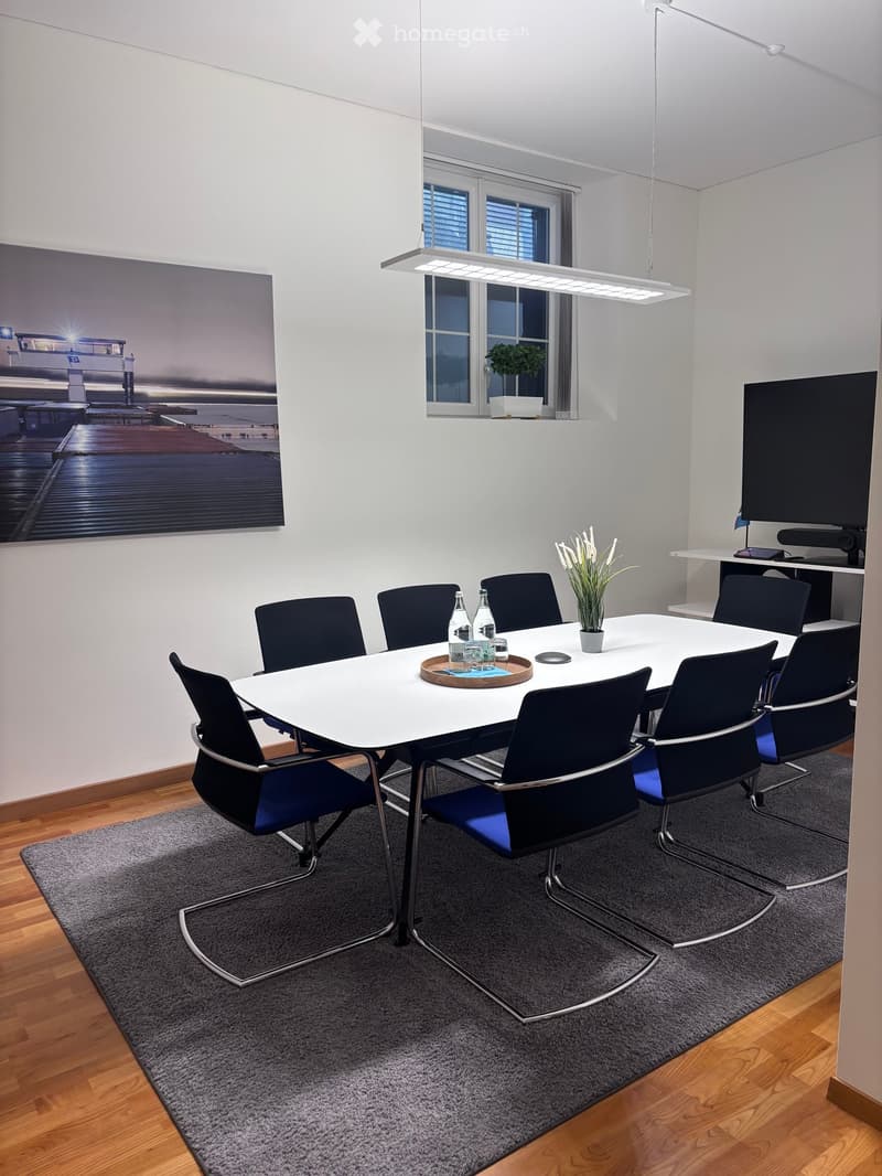 A modern meeting room with a white table, blue chairs, a tray, and a plant. A television is mounted on the wall, and a shelf is placed below it.