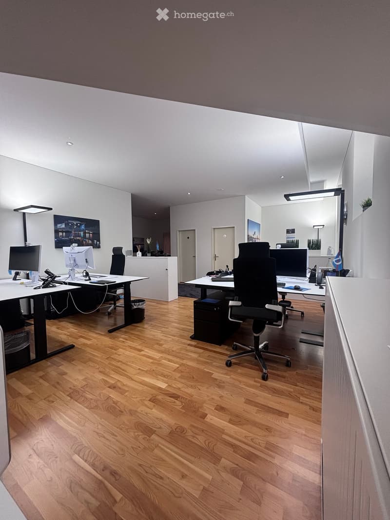 Modern office with wooden floors, white walls, and two desks equipped with computers, chairs, and various office equipment.