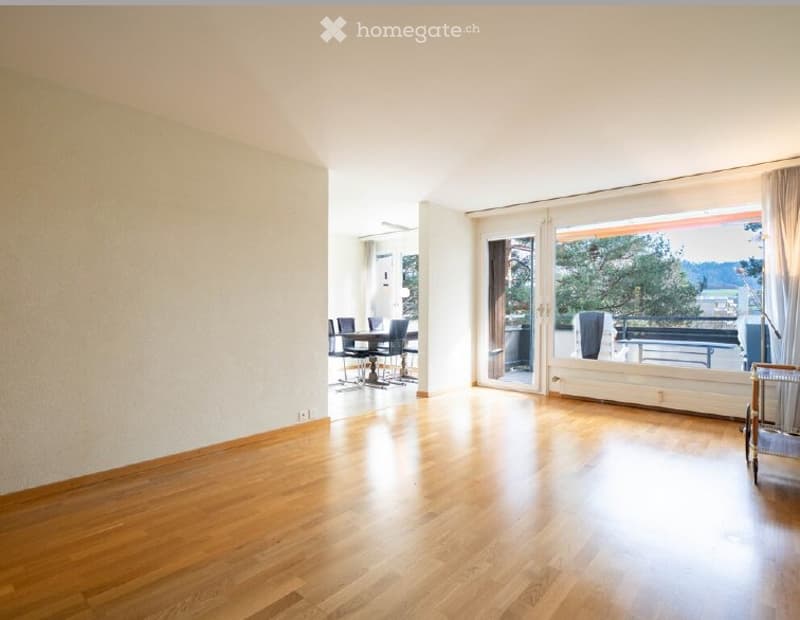 Bright, open living space with wooden flooring, large sliding glass doors leading to a balcony, and a small dining area with a table and chairs.