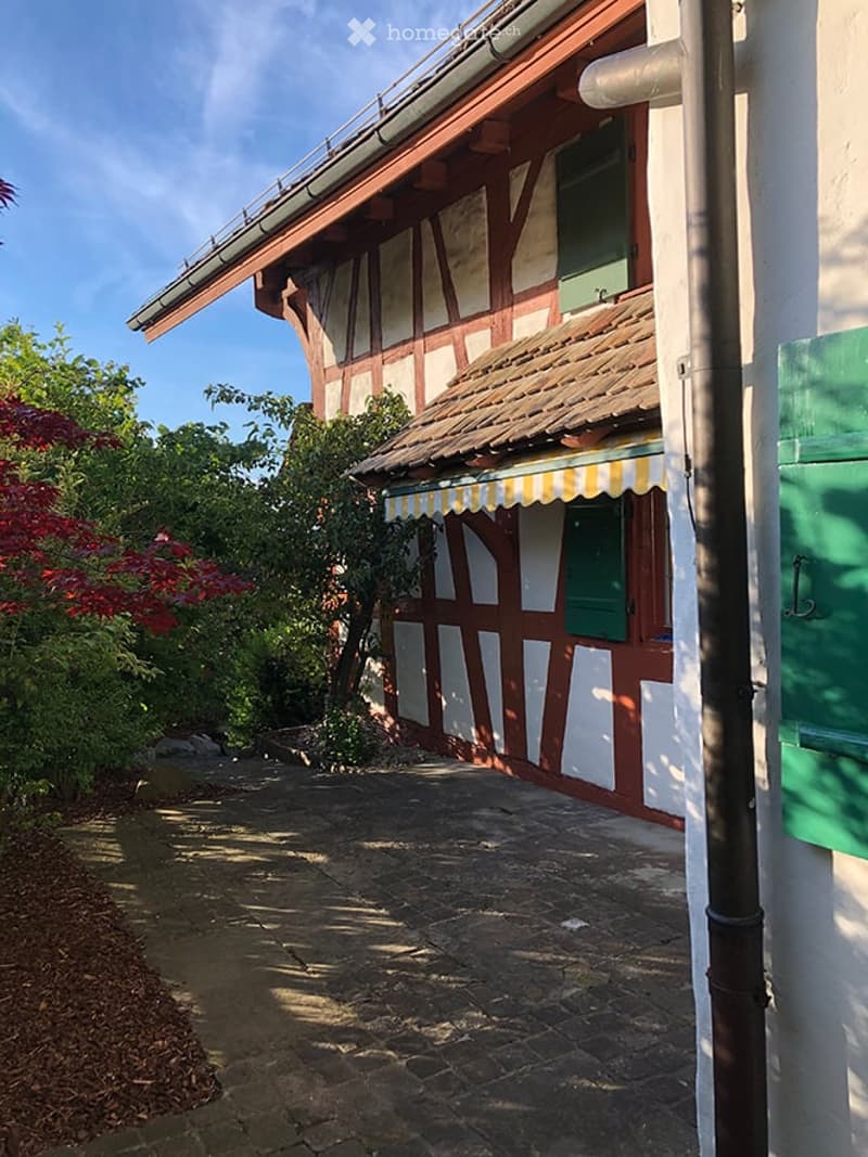 Traditional half-timbered building with white and red wooden beams, green shutters, and a tiled roof, surrounded by greenery and a stone pathway.