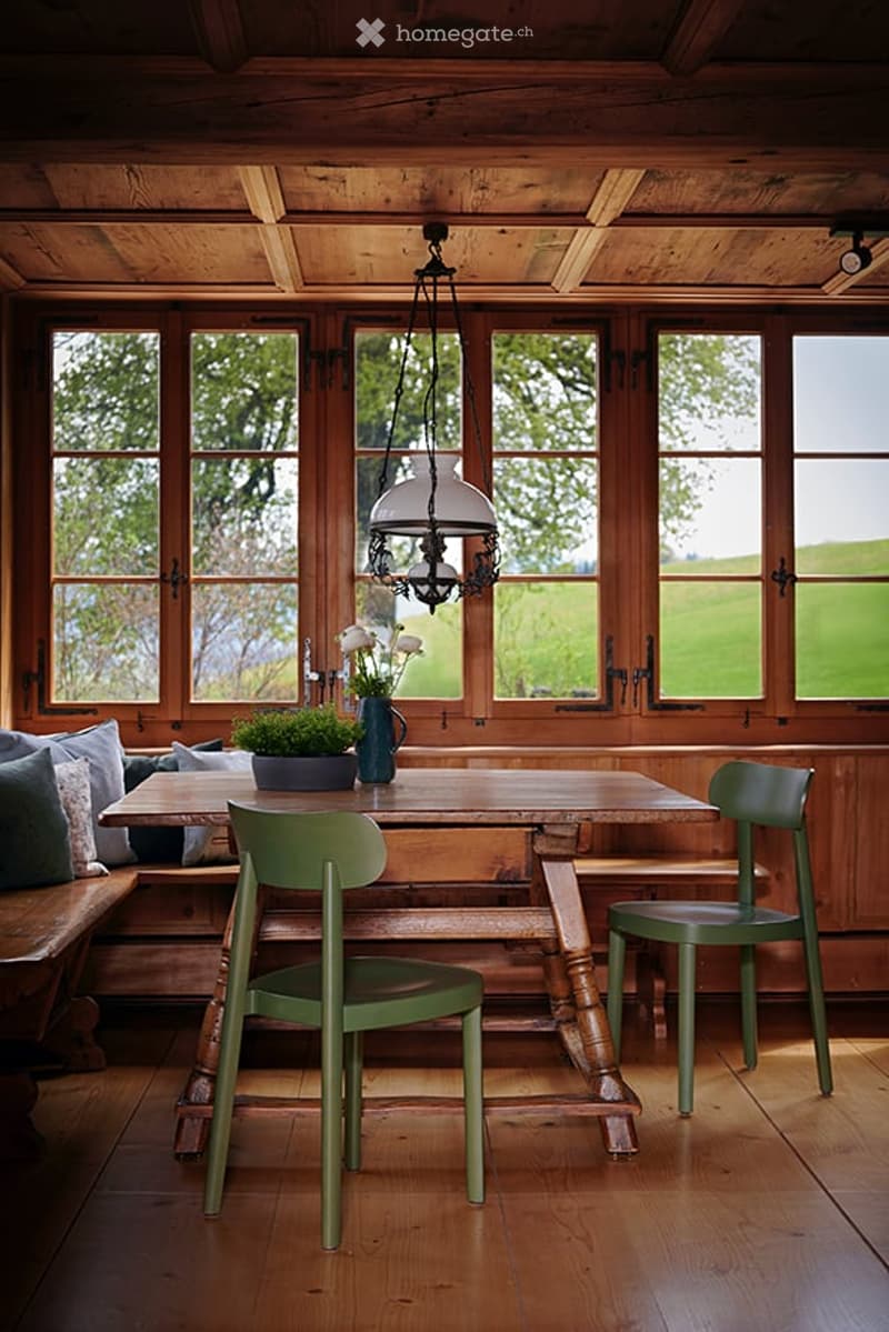 A rustic dining area with wooden table, green chairs, bench seating, and a hanging lantern light fixture. The room features large windows with a view of greenery outside, and a vase with white flowers on the table.