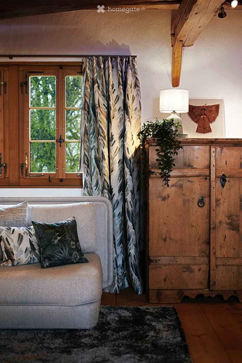 A cozy living room with wooden beams, a large window with patterned curtains, a grey sofa with decorative pillows, a wooden cabinet with a table lamp and plant, and a dark area rug on the floor.