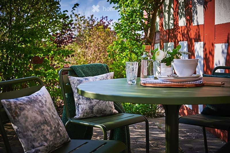 A well-maintained outdoor seating area with a round metal table, four green chairs with patterned cushions, a glass, a bottle, and a small flower arrangement on the table.
