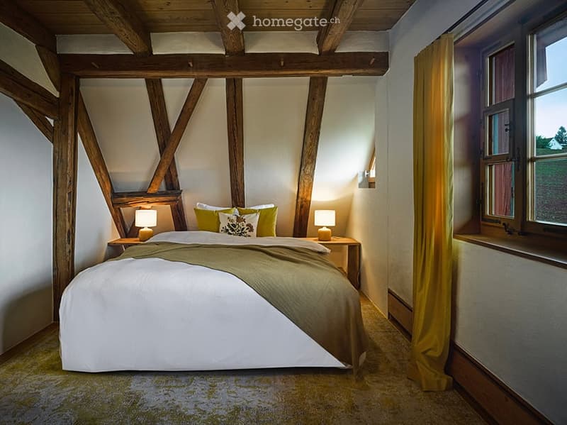 Cozy bedroom with exposed wooden beams, a large bed with green and white bedding, nightstands with lamps, and a window with yellow curtains.