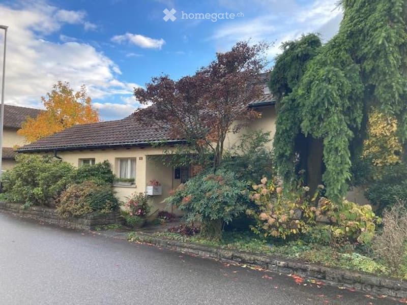 Single-family house, beige exterior, brown roof, two windows, small porch, surrounded by trees and shrubs