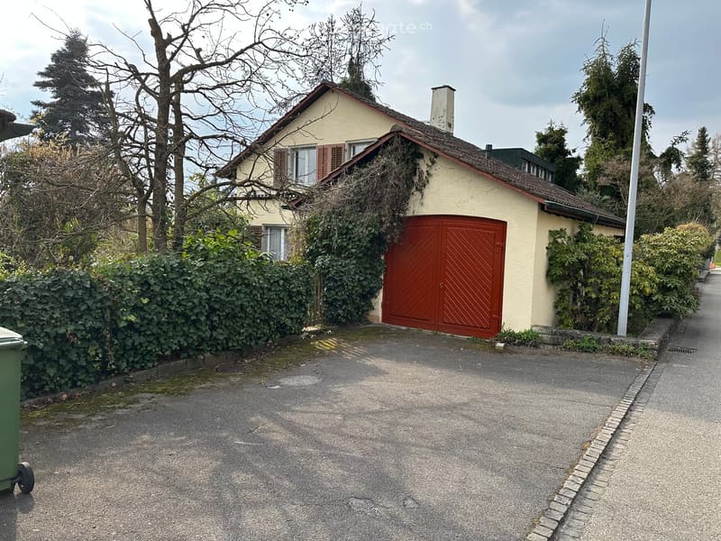 2 story house, beige walls, red garage door, chimney, surrounded by trees and bushes