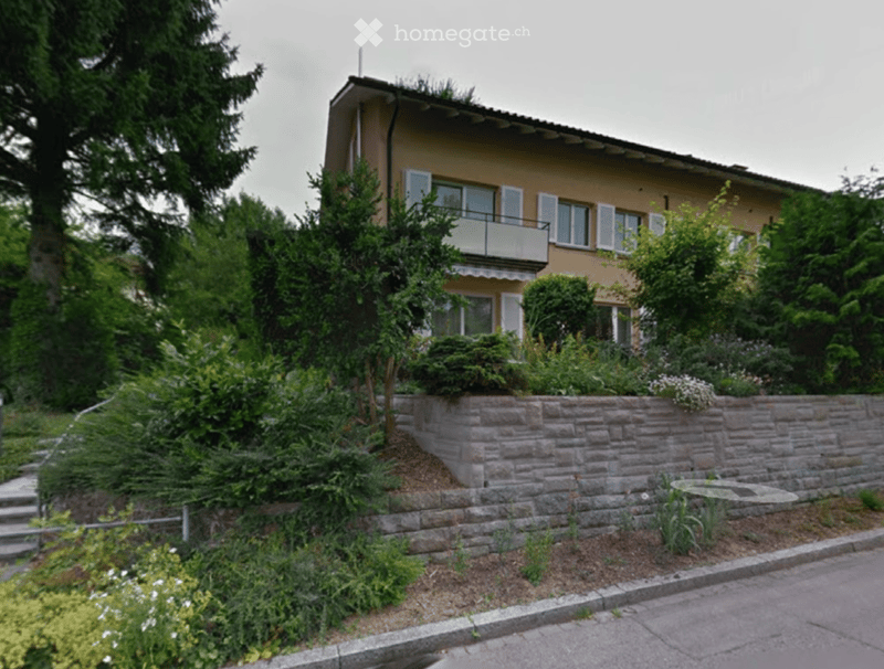 Two-story house with beige exterior, white-framed windows, and a balcony on the upper floor. The house is surrounded by lush greenery and a stone retaining wall.