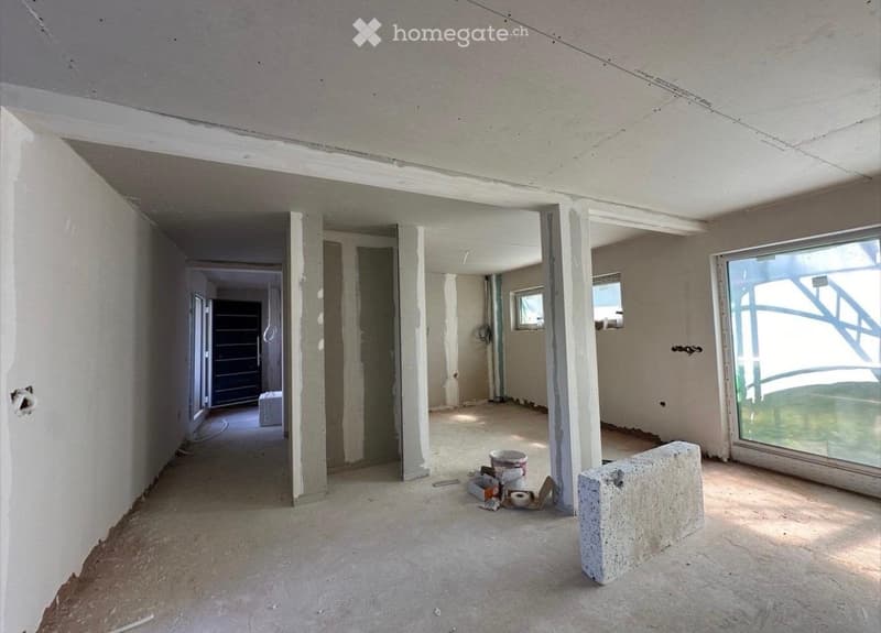Empty room with unfinished concrete walls and floor, visible construction materials and tools, large window on one side, and open doorway leading to another room.