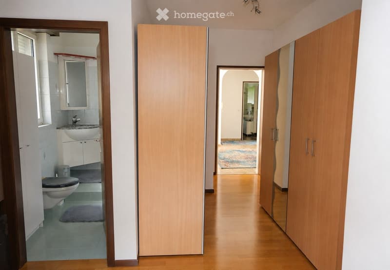 A well-lit hallway with wooden flooring, leading to a bathroom and another room. The bathroom features a toilet, sink, and mirror, while the adjacent room has a colorful rug and a glimpse of furniture.