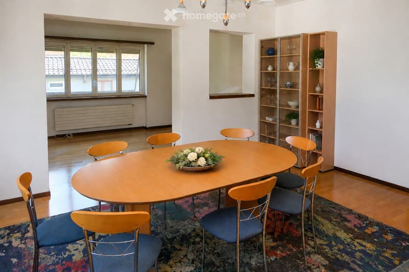 A well-lit dining area with a round wooden table surrounded by six chairs, a bookshelf with decorative items, and a colorful rug on the floor.