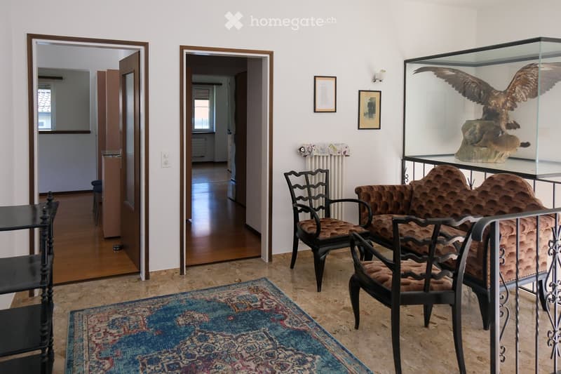 Living room with a patterned rug, a sofa, chairs, a small table, and a display case containing a bird sculpture. The room has wooden flooring and white walls with framed pictures.