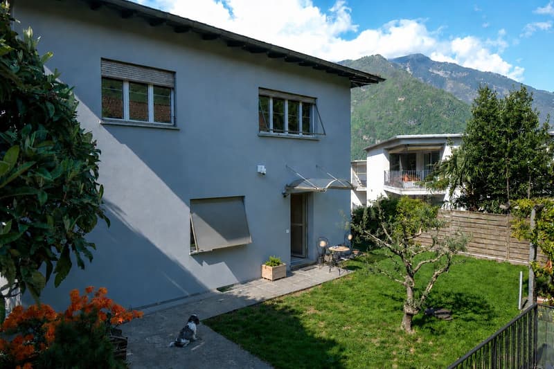 Two-story house with light blue exterior, multiple windows, and a small patio area with outdoor furniture. The surrounding area includes a well-maintained garden with a tree, flowers, and a dog sitting on the pathway.