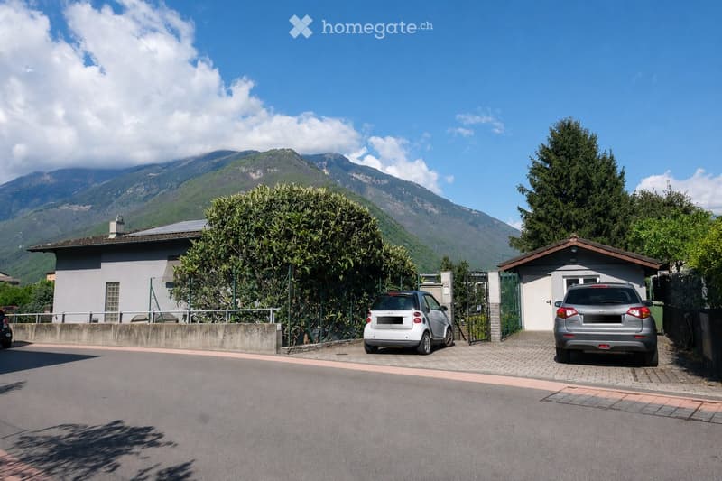 Two-story house with a garage, surrounded by greenery and mountains in the background. Two cars are parked in front of the house.