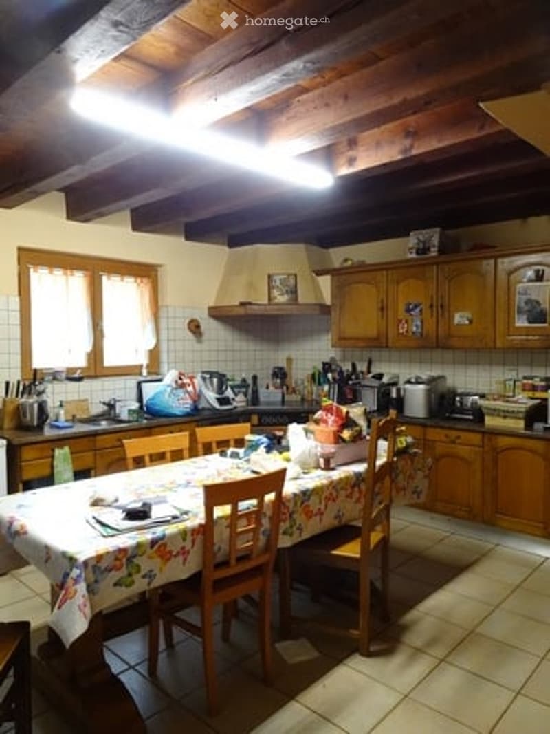 A cozy kitchen with wooden cabinets, a dining table with chairs, and various appliances including an oven, fridge, and microwave. The room features exposed wooden beams on the ceiling and a tiled floor.
